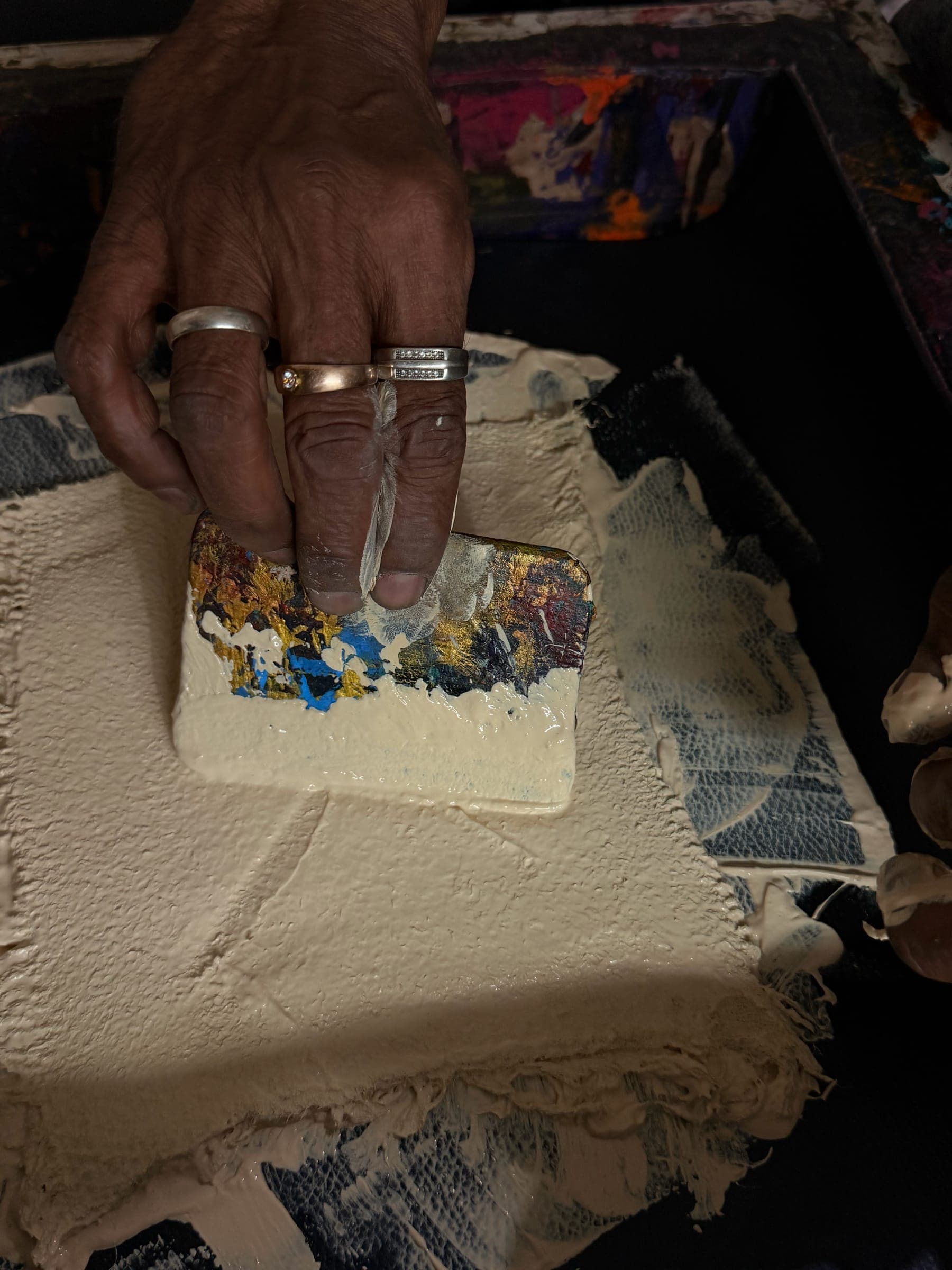 A ringed hand rests a carved block on a pad of thick printing paste