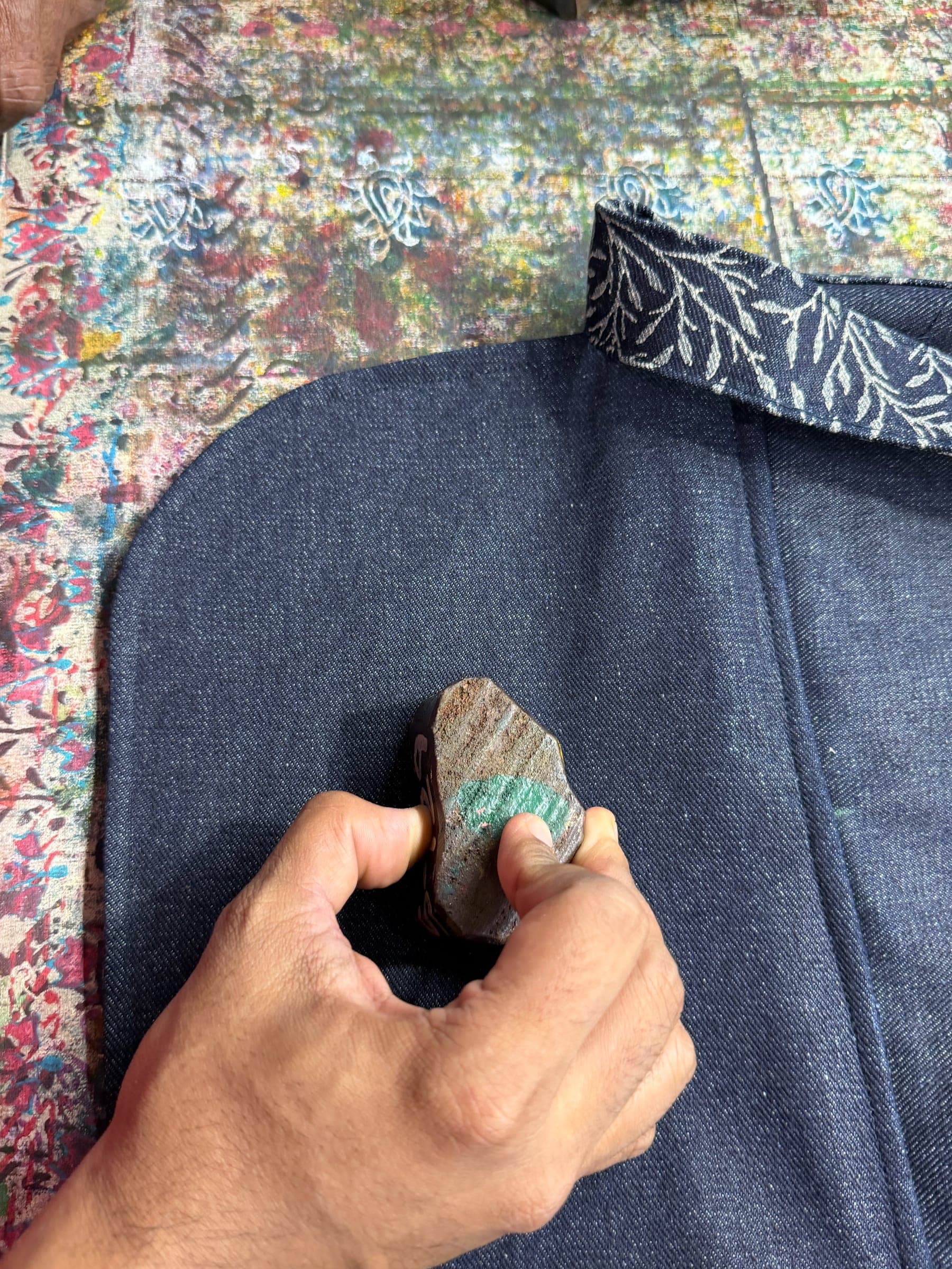 A maker's hand presses a small carved block onto a denim bag, its block-printed vine strap laid alongside