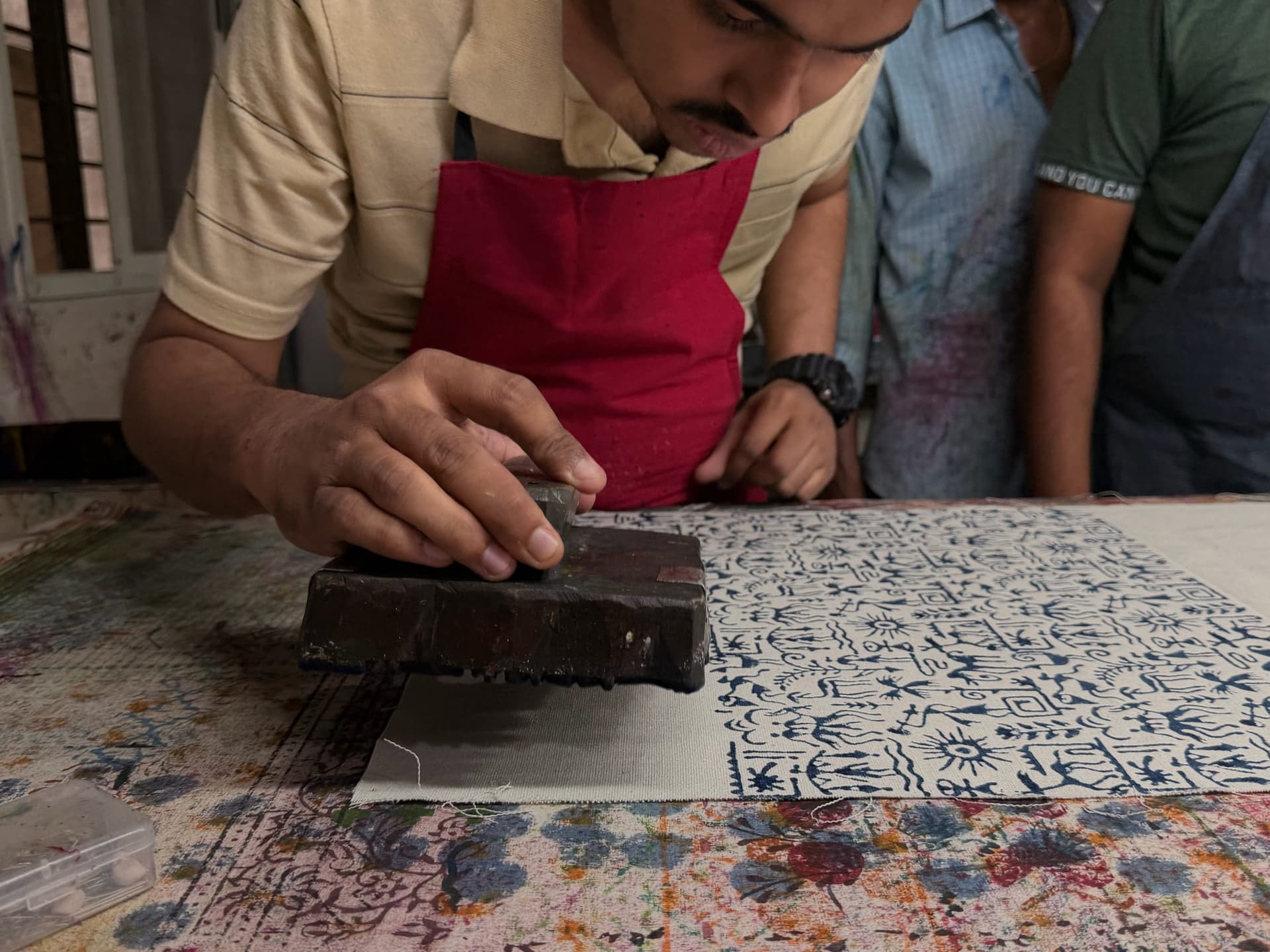 A maker leans in close to press a long carved border block onto fabric, colleagues watching the row land